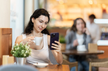 Young woman drinking hot black coffee and looking at phone screen, scrolling through social media feed and chatting with friends