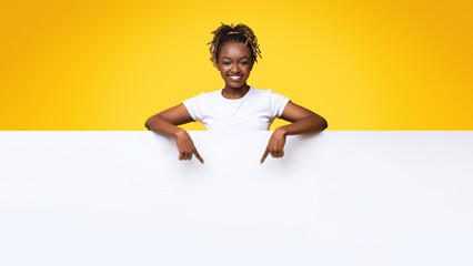 Smiling black woman pointing at white empty board for advertisement over yellow background. Cheerful african lady leaning on big blank advertisement placard, indicating at empty space for text