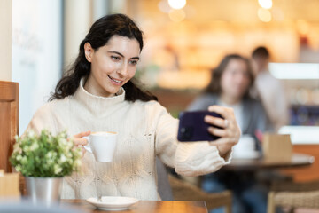 In cozy cafe, girl client with phone drinks coffee. Visitor chats in messengers, makes purchases on online marketplace.