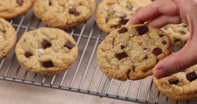 Chocolate chip cookies freshly baked topped with flaky salt on a cooling rack, woman hand taking a cookie