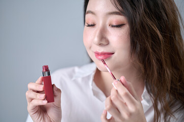 Obraz premium A woman with long hair, wearing a white shirt, is happily applying lip gloss while doing her makeup