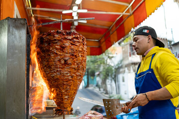 Street vendor slicing meat from a rotating trompo, or vertical spit, to prepare tacos al pastor at...