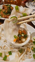 Dumpling held by chopsticks above dipping sauce and other cuisine. Dumpling has garnish on top....
