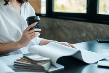 Professional woman in a white shirt holding a coffee cup while reviewing documents at a modern office desk.