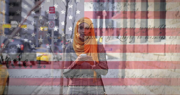Image of american flag waving and constitution text over biracial woman in hijab using smartphone - Powered by Adobe