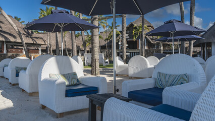 White wicker armchairs with soft cushions are placed on the sandy embankment. Small wooden tables. Sun umbrellas. The thatched-roof hotel buildings are in the distance. Palm trees against the blue sky