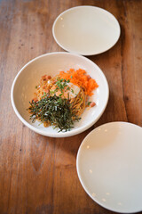 bowl of spaghetti topped with shrimp, roasted seaweed, and eggs is beautifully presented on wooden table, accompanied by two empty white plates, creating cozy dining atmosphere