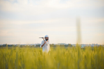 A farmer in traditional white clothing stands in a lush wheat field, holding a pickaxe on his shoulder with a distant village in the background.