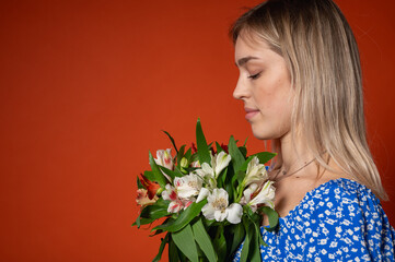 Naklejka premium Smiling, pretty young girl in a blue floral dress holding a fresh colorful bouquet of flowers, posing on a bright orange background. Spring mood, natural beauty, cheerful mood, positive