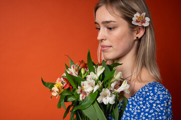 Naklejka premium Smiling, pretty young girl in a blue floral dress holding a fresh colorful bouquet of flowers, posing on a bright orange background. Spring mood, natural beauty, cheerful mood, positive