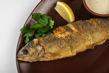 Crispy fried whole fish served on a brown plate with fresh parsley, lemon wedge and creamy sauce. A...