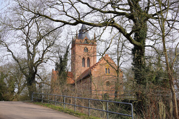 A brick Gothic church with a high tower and spire, partly hidden by leafless trees.