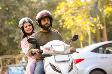 young indian couple riding on scooter wearing helmets in the city streets. husband and wife enjoy...