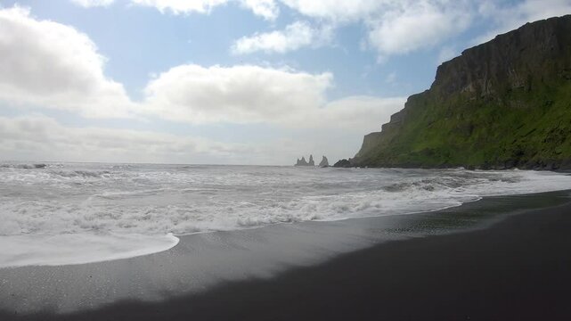 Waves crash onto the black sand beach of Reynisfjara in Vik, Iceland, in slow motion, with rugged basalt formations and the Atlantic Ocean creating a dramatic coastal scene.
