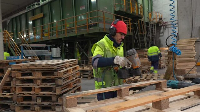 Skilled carpenter assembles wooden pallets using a pneumatic nail gun in a bustling recycling plant