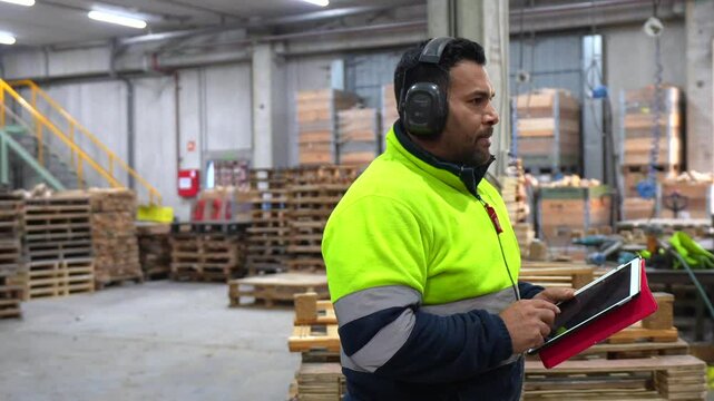 Factory supervisor wearing safety headphones using a tablet and checking wooden pallets in a recycling plant