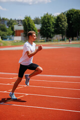 Fototapeta premium Young male athlete sprinting on a red running track, wearing sportswear, training outdoors on a sunny day, focused on speed and performance.