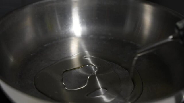 A bottle of oil is poured into a pan. The pan is silver and has a dark spot on it