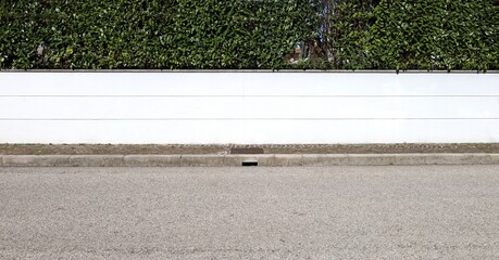 White plaster fence with horizontal stripes and  hedge on top. Sidewalk and urban street in front....