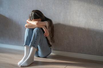 A woman sits on the floor, hugging her knees tightly while appearing lost in thought, displaying...