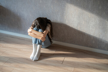 A woman sits on the floor, hugging her knees tightly while appearing lost in thought, displaying...