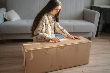In a cozy living room, a woman sits on the floor unpacking a large cardboard box while sitting near...