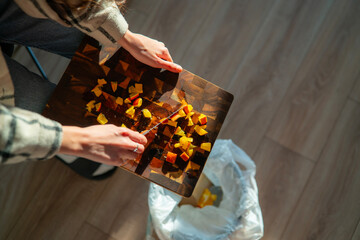 Individual prepares vegetables on a cutting board, efficiently wiping peelings and scraps directly into a waste bin nearby.