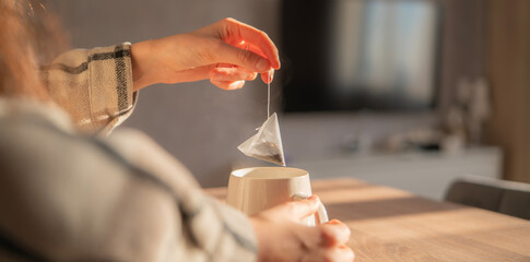 Hand holds a tea bag above a white cup as sunlight streams into a cozy home, creating a warm and...