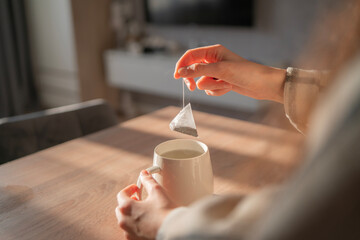 Hand holds a tea bag above a white cup as sunlight streams into a cozy home, creating a warm and...