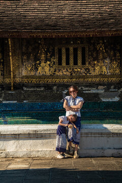 Happy mother and daughter in traditional Lao attire sitting together in front of a golden stenciled temple wall in Luang Prabang, Laos.