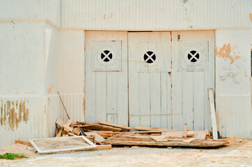 Weathered wooden doors in shades of faded blue, green, and white with circular windows and peeling paint stand out against a textured white wall. Vibrant hues and scattered wooden debris create a dyna