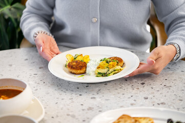 A white plate with gourmet breaded patties, creamy sauce, roasted potatoes, and greens, elegantly plated. A bowl of soup is on the table, held by a person in a gray sweater.