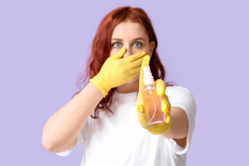 Beautiful young shocked woman in rubber gloves with bottle of sanitizer on purple background
