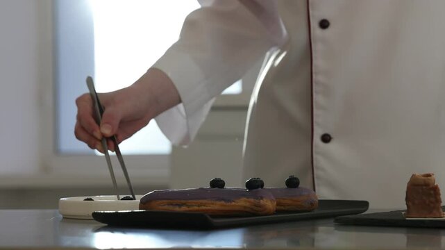 Confectioner decorating eclairs with blueberries at table indoors, closeup