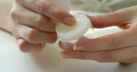 Woman opening and taking moisturizing lip balm from jar at white table, closeup