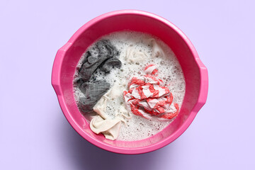Plastic basin with dirty laundry, foam and water on lilac background