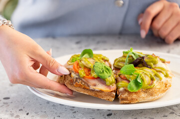 A close-up of a hand holding bruschetta with tomatoes, cheese, green sauce, and basil on a white plate, set against a gray countertop background. Perfect for culinary and food themes.