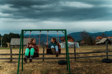 Two young girls enjoy swinging on a swing set in a countryside setting, their laughter echoing against the backdrop of a dramatic, cloud filled sky