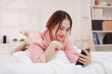 Asian woman using mobile phone to play social media on bed, smiling happily at home