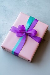 A pink gift box with purple ribbon and green ribbon. The ribbon is tied in a bow. The box is sitting on a grey surface