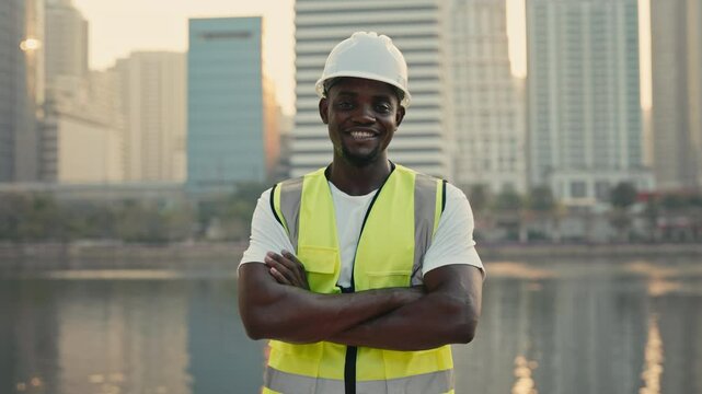 Portrait of confident African American builder crossing arms standing on megapolis embankment. Male professional construction engineer with protective clothing against skyscrapers