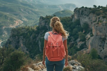 Naklejka premium A young Caucasian woman stands on a rocky outcrop, gazing at the breathtaking mountain view with a pink backpack.