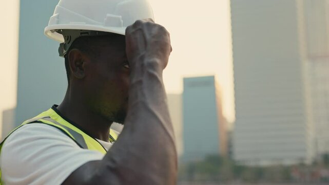 Portrait of positive African American builder putting on white helmet standing against multistorey buildings. Man constriction employee shows safety supply in city. Builder job