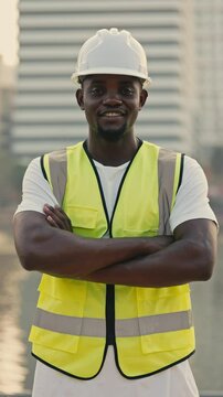 African American man in hard hat stands with arms crossed against urban building. Male builder with beard in reflective vest radiates professionalism looking at camera. Vertical shot