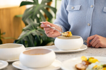 A close-up of a person in a light blue sweater holding a spoonful of food above a white bowl on a table, surrounded by dishes of colorful meals and a green plant background.