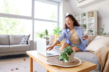 Young Asian woman using laptop and tablet computer on grey sofa at home