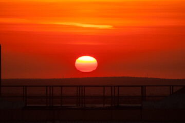 Fototapeta premium View from the roof of a multi-storey brick house in the early morning or late evening to a beautiful bright orange crimson red sunrise or sunset of the heavenly sun from under the hill on which trees.