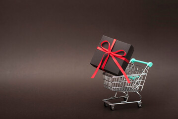 A small metal shopping cart holding a black gift box tied with a red ribbon. The cart is placed on...