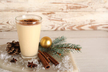 Glass of tasty eggnog cocktail with star anise, cinnamon, fir branch and Christmas ball on white wooden background