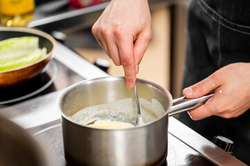 Close-up of a person stirring creamy sauce in a stainless steel pot on a stovetop, wearing a dark apron. Background includes a pan with lettuce. Food preparation scene.
